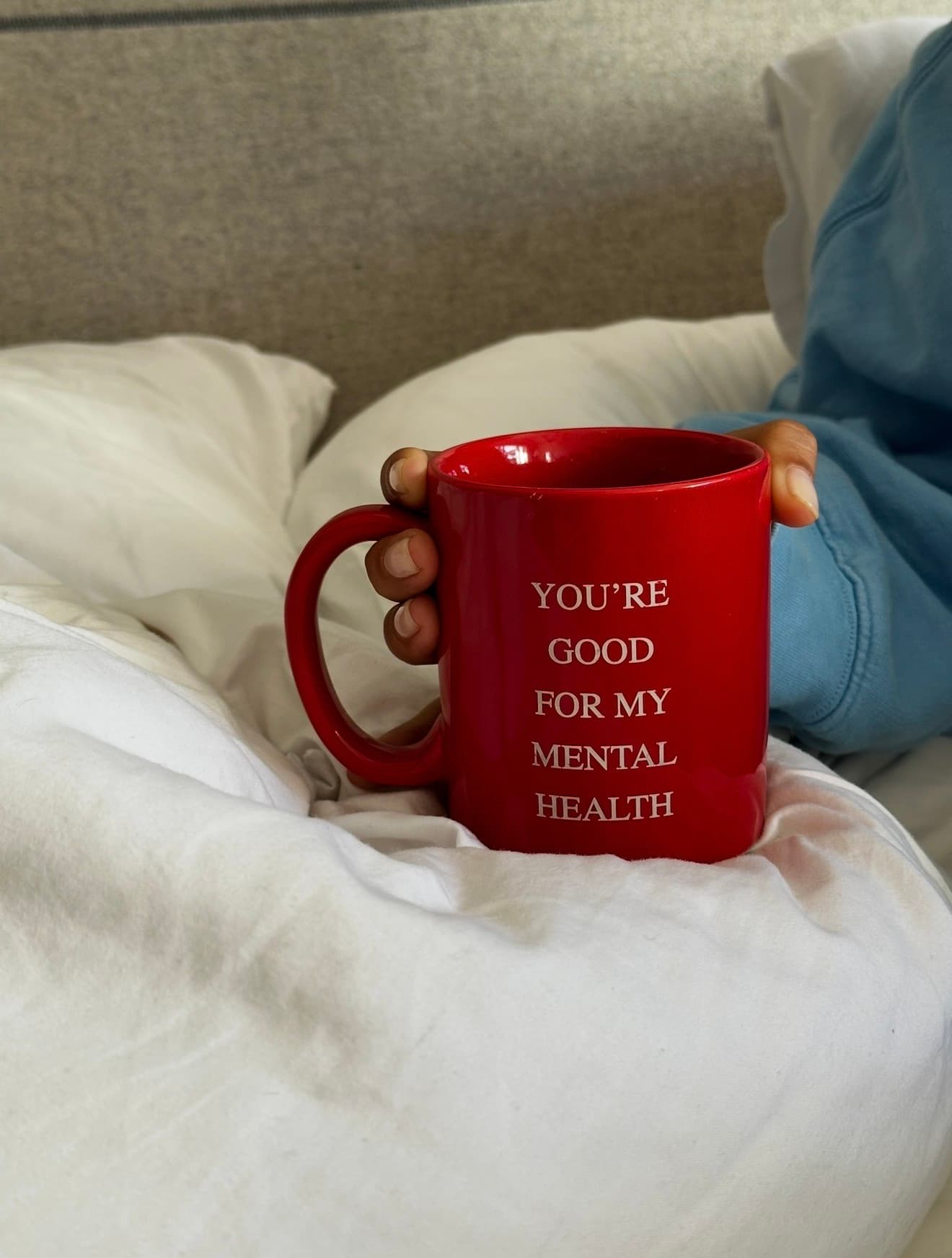 A red mug reading You're Good For My Mental Health held in bed